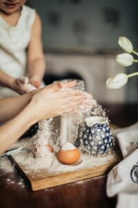 Hands of parent and child baking together, scattering flour on a wooden board with eggs and teapot nearby.