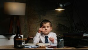 A young boy in a shirt and tie sitting at a desk with office supplies, under dramatic lighting.