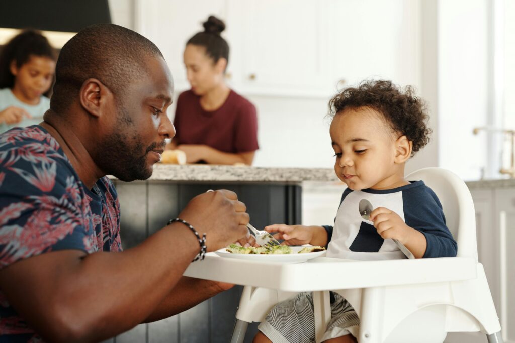 A father feeds his son in a high chair while family shares a meal in the kitchen.