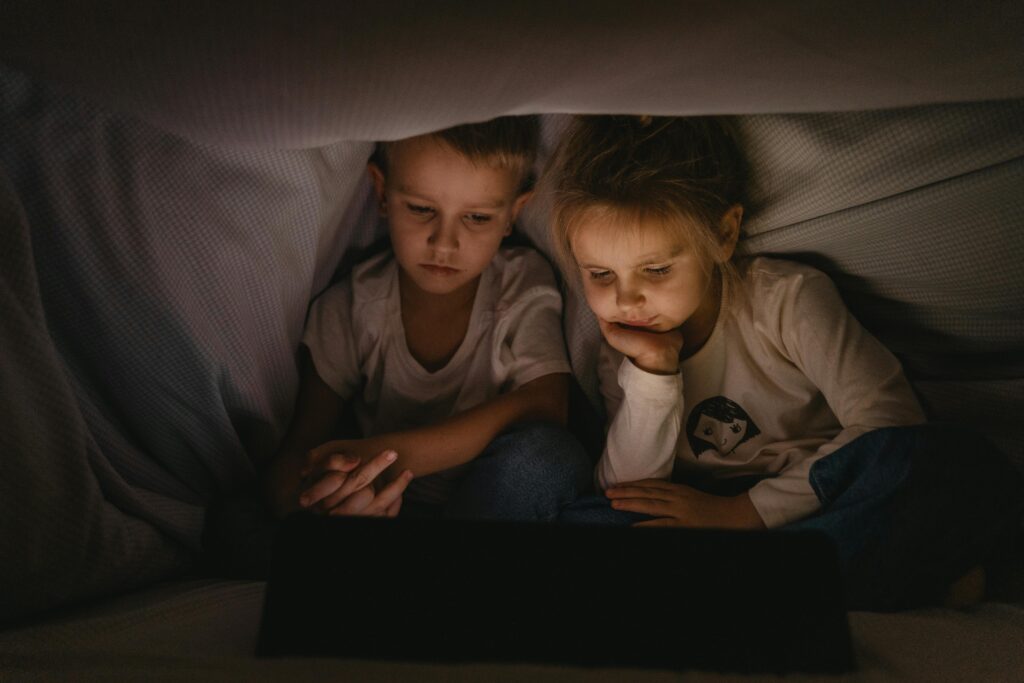 Two young children watching a tablet under a blanket, creating a cozy nighttime atmosphere.