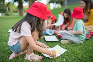 Children engaged in outdoor drawing activities on a sunny day wearing red bucket hats.